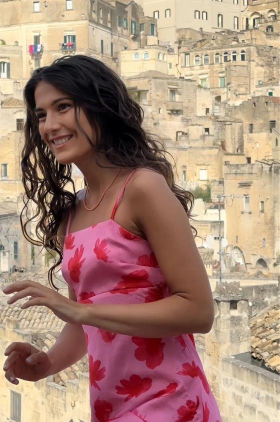 "Donna con capelli lunghi e ricci che indossa un abito rosa con motivi floreali rossi, in piedi davanti a uno sfondo di antichi edifici in pietra e cielo sereno."